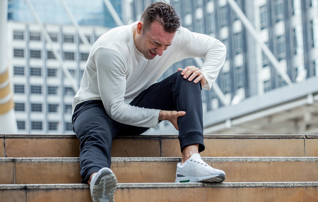 Mężczyzna siedzi na schodach na zewnątrz, ściskając nogę z bólu, z wyrazem bólu na twarzy. Ma na sobie białą koszulę z długim rękawem, czarne spodnie i białe trampki.