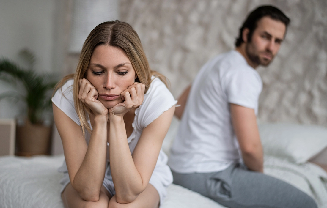 Kobieta siedzi na łóżku, wygląda na smutną i zamyśloną, z brodą opartą na rękach, podczas gdy mężczyzna siedzi za nią, odwrócony tyłem. Oboje wyglądają na zdenerwowanych.